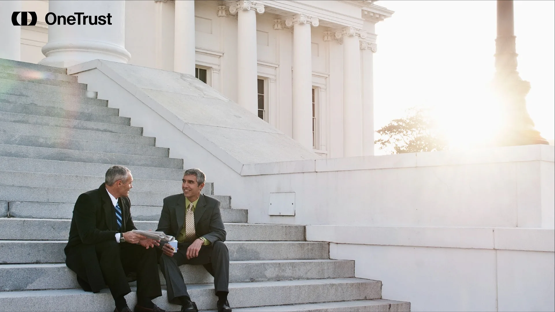 Two men in suits sit on the steps outside a neoclassical government building at sunset.