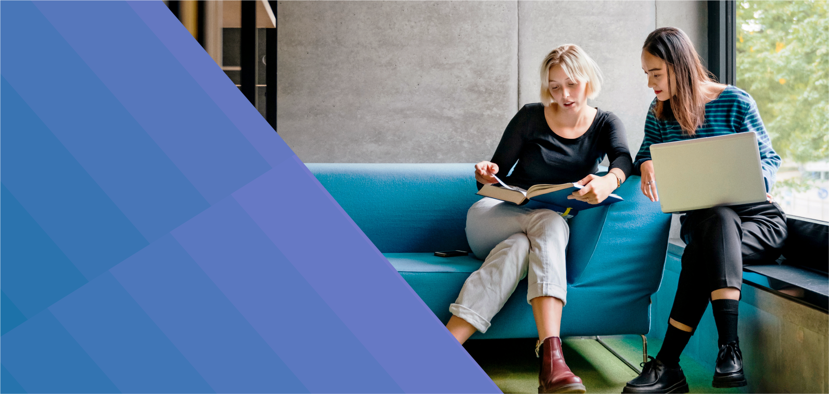 photo of a white woman and an Asian woman in their 20s collaborating on a project. One woman is sitting on a blue couch white the other sits on a low windowsill. 